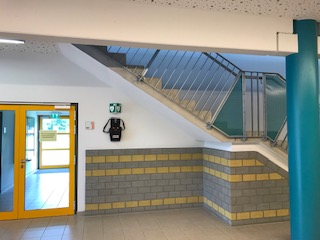 AED wandmontiert unter der Treppe, rechts von der Glastür zum Sportplatz